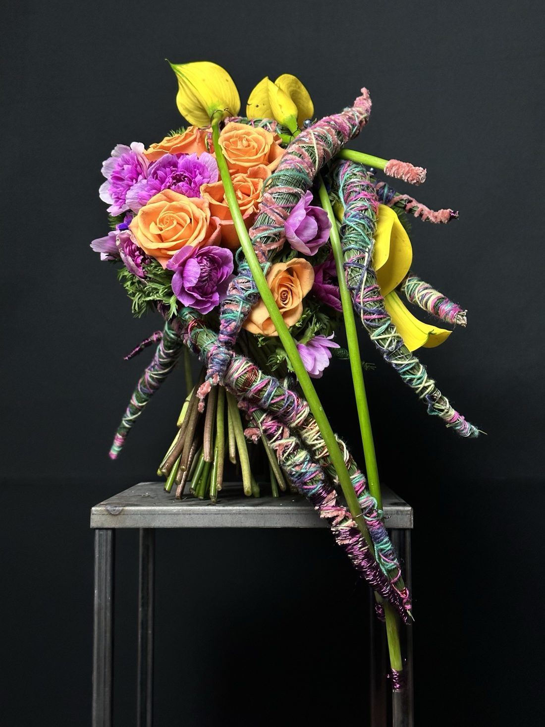 Mazzo legato di Mario Garofalo con rose arancioni, anemoni viola e calle gialle, attraversato da elementi lunghi avvolti in filati colorati.